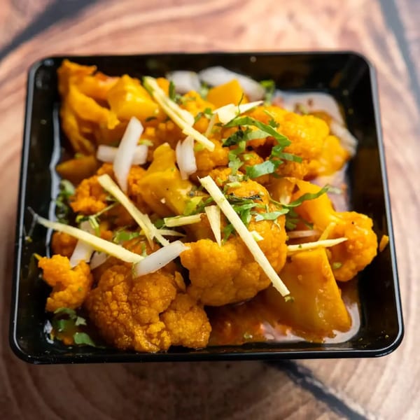A black bowl filled with orange cauliflower and potatoes, garnished with sliced onions, ginger, and cilantro.