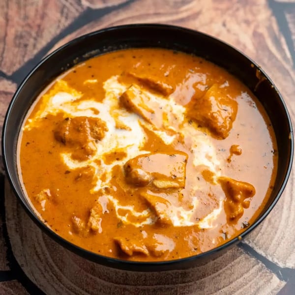 A black bowl filled with rich orange butter chicken, swirled with white cream, on a wooden surface.