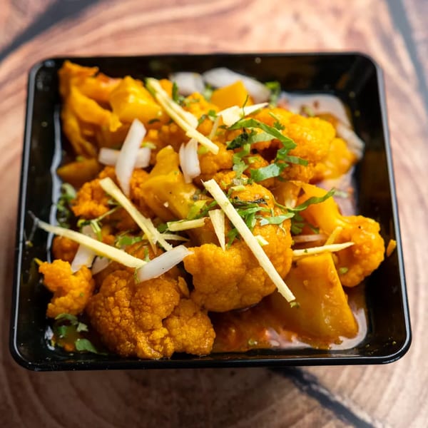 A black square dish filled with orange cauliflower and potatoes, garnished with onions, ginger, and cilantro.