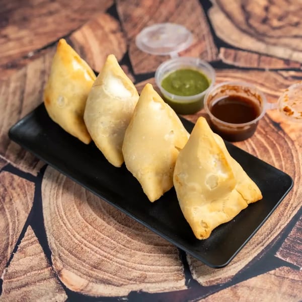 Four golden samosas on a black plate with green and brown dipping sauces on a wooden surface.