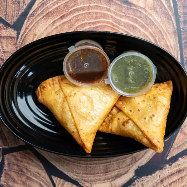 Two golden-brown samosas and two small containers of chutney on a black plate.
