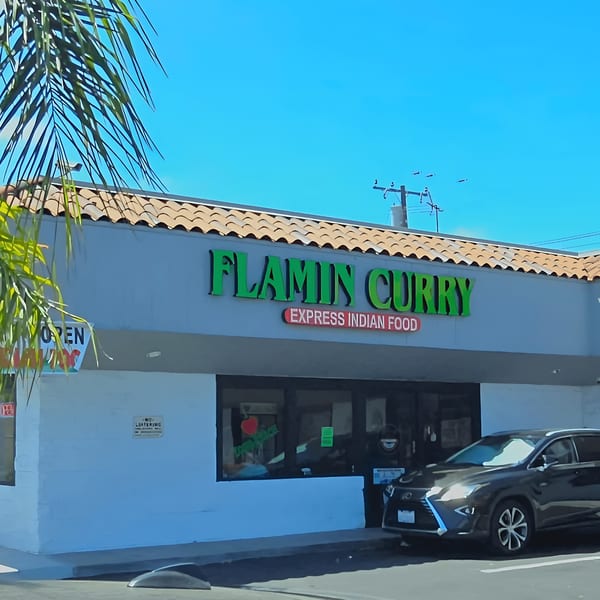 Exterior of Flamin Curry, an Indian restaurant, with a black car parked outside under a blue sky.
